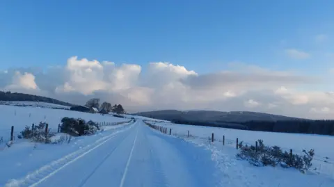 Buckbeaks Flight/BBC Weather Watchers A rural road is covered in snow. Deep snow also covers surrounding fields and the roof of a farm in the distance. It is daytime and there are clouds in the sky.