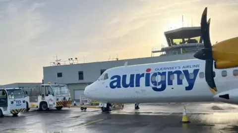 An Aurigny aircraft sits on the tarmac outside a hanger  