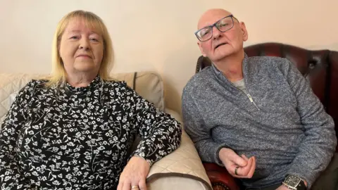 BBC A photo of Florence and Peter sitting a living room. Florence is on a sofa with her arm on the arm rest. Peter is in a leather armchair and is leaning slightly to get in closer to Florence in the photo. He is bald with glasses and grey zipper sweater. She has shoulder-length blonde hair and floral dress.