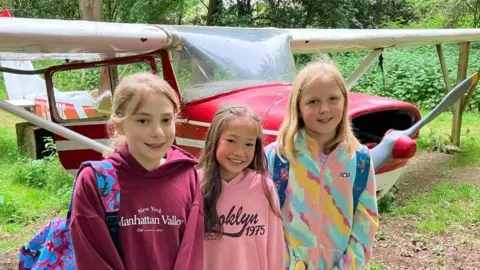James Grant/BBC Three young girls in hoodies and coats stand by a red and white plane smiling at the camera.