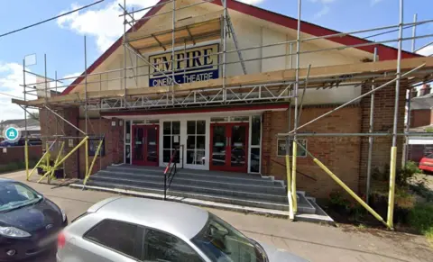 A one-storey theatre building with scaffolding and glass doors with steps leading to the entrance