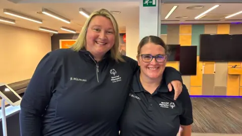 A woman with blonde hair on the left has an arm around the left shoulder of the woman on the right who is wearing glasses. They are both wearing black tops and smiling at the camera. 
