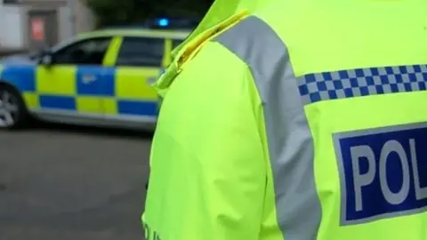 BBC Police officer with his back to camera, facing a police car