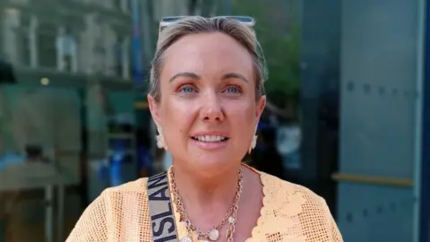 A woman, with blonde hair tied up, wearing a light orange top. 