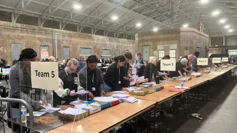 An election count with a table and people behind it sorting ballot papers into piles.