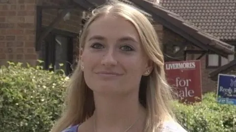 A blonde woman standing in front of a row of houses with for sale signs out the front.