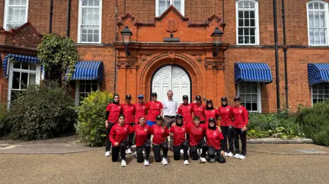 Tom Williams/BBC Sir Laurie Bristow poses with two rows of the women's cricket team, dressed in black trousers and red T-Shirts. The front row of women kneel. They stand in front of Hughes Hall, which has a white arched door and red brick attractive building. There are blue awnings over some of the windows.