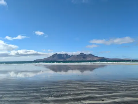 Helen Maclean A wide sandy beach with shallow ripples in the sand, reflecting the blue sky and clouds, with an island visible across the water.