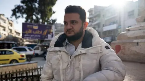 Lee Durant/BBC A bearded young man in a white coat stands by a road in a residential area of ​​Idlib