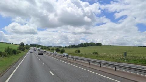 A Google Street View image of the A34 between East IIsley and Chieveley. Green fields are on either side with trees in the distance, and a blue sky overhead.