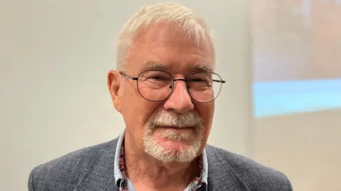 Peter wearing a grey blazer and a blue shirt with a checked border on the collar 
