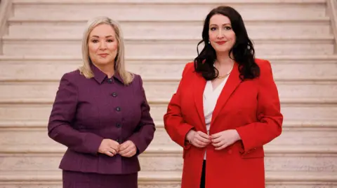 PA Media Michelle ONeill - who has shoulder-length blonde hair- in a purple suit and Emma Little Pengelly - who has longer dark brown hair - in a red suit with a white blouse visible under her jacket. They are standing in front of a set of marble stairs in the Great Hall at Stormont.