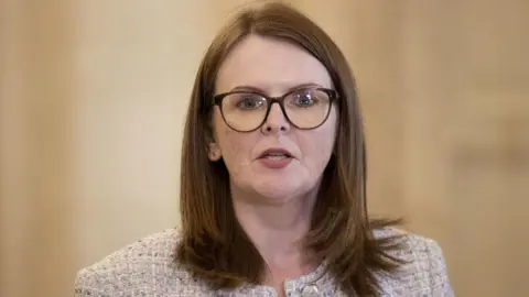 PA Media Caoimhe Archibald in glasses at a broadcast microphone. She is wearing glasses and textured blazer, and is in the middle of speaking