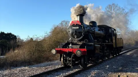 Jo Morris A black British Steam locomotive - 78019 with red NVR sing on top. Black-greyish steam can be seen coming out of its chimney. It is riding along train tracks with trees in the background. 