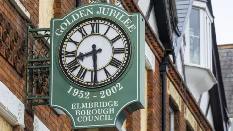 A Golden Jubilee commemorative clock on the wall of a red brick building. It is dark green in colour and has the name of the council on it and the dates 1952-2002.