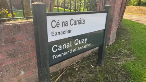 A street sign reading 'Canal Quay - Townland of Annagher' in English and in Irish. The sign is pitched in grass and there is a red brick wall behind it. 