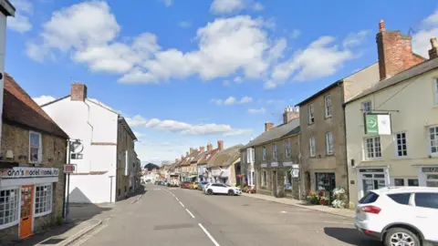 Google A view down Wincanton high street: a road with two-storey quaint buildings like cottages each side, some shops, cafes and pubs. A few cars are parked down the right-hand side. It is a sunny day with a blue sky.