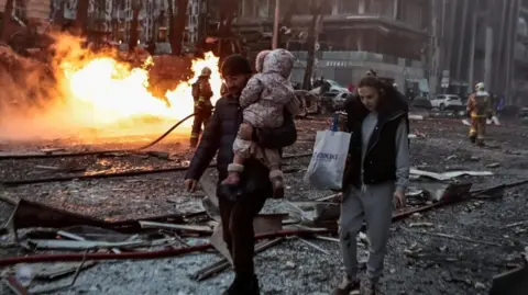 Reuters A man holds a small child as a fire burns after a Russian strike in Kyiv, Ukraine. Photo: 20 December 2024