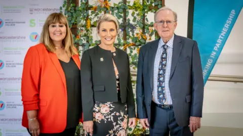 National Star Woman in a red blazer and black top is standing next to woman in a pink floral dress and cropped black blazer. There is a man in a light grey suit and check-shirt standing on the end