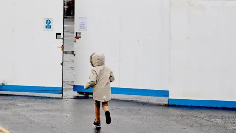 A child running into a screen put up in front of a hotel. They are wearing a long cream coat and brown trousers. 