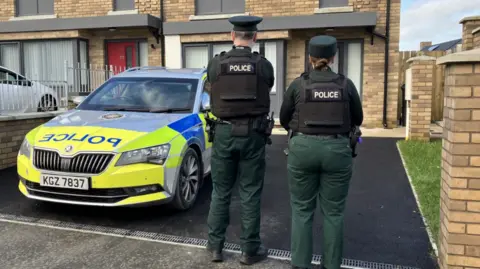 Show a police car in yellow, light green blue and grey with the PSNI cresh at the word 'police' on the bonnet. Two police officers, a male and female, in green uniforms and black flak jackets with the word 'police' stand to the right of the car in the driveway of a house, facing the house. 