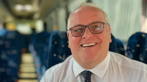Charles Sanders is wearing a white shirt and blue tie, which has brown and white dots on it. He is sitting in a passenger seat on a bus. He is also wearing glasses.