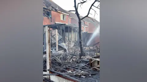 Embers and burnt struts belonging to a porch-type structure at the back of a house. There is a blackened tree and smoke is emanating from the ground. Broken glass and debris lie on the ground along with burnt fence panels. 