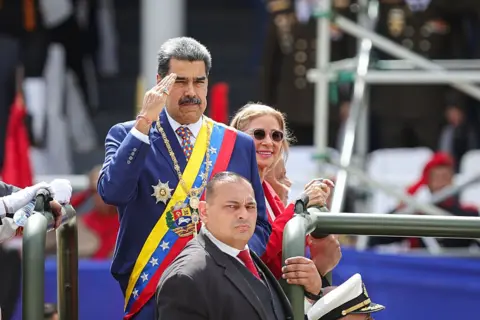 Getty Images President of Venezuela Nicolas Maduro, wearing a suit and military sash, salutes as he arrives for a military parade as part of 214th anniversary of Venezuela's independence celebrations on July 5, 2025 in Caracas, Venezuela. A man stands in front o fhim holding a bar and a woman smiles behind him.
