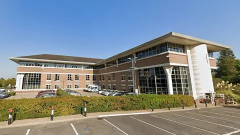 External shot of Esher Civic Centre which is a large modern building in a pale brick with lots of glass windows and car parking around it. There are low shrubs around the car park and trees are seen behind it.