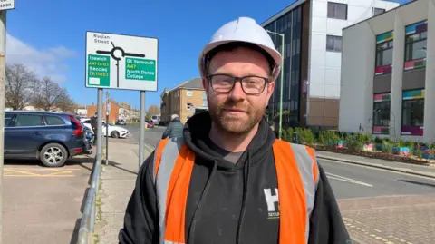 Vikki Irwin/BBC Ryan Watling is wearing a high-visibility orange gilet over a hooded sweatshirt. He has a white hard hat on and is wearing glasses. He is standing on the pavement by a car park. 