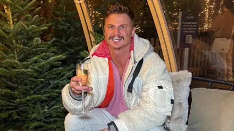 Tomasz Wisniewsk Tomasz Wisniewski sat on a chair in front of Christmas tree with a glass of champagne wearing a white coat.