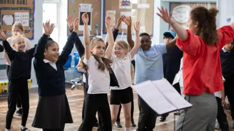A woman in a red shirt stands waving her hands in the air whilst a number of young children in navy school uniforms face her.