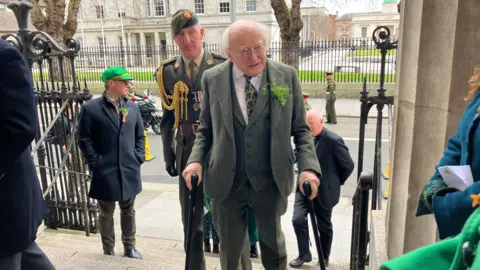 Michael D Higgins pictured waking through the door of a stone church. He is wearing a green suit with a bunch of shamrock in his lapel. He is holding two walking sticks. A man in military dress is walking behind him.