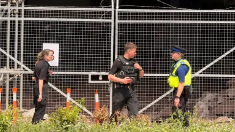 Police stand near silver gates with silver wire above the gates. A woman is in black on the left and two men, including one in a yellow hi-vis jacket, are on the right of the photo.