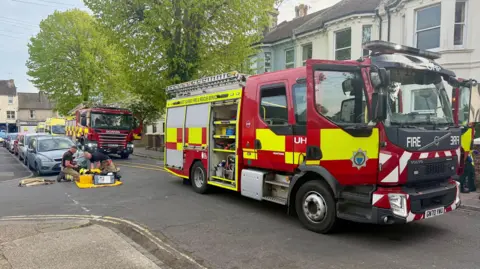 Two fire engines parked in a residential road. Two firefighters are on their knees next to equipment on the floor.