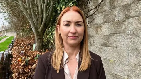 Alison Lowery has long red hair and is wearing a brown jacket and white blouse