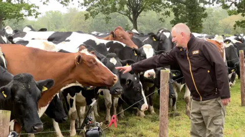 Tom Kimber is reaching out to pet one of a row of cows behind a wire fence. He is wearing a black jacket with a yellow zip and khaki combat trousers. He has short white hair.