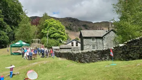 Langdale CofE Primary School in the Lake District, Cumbria. The school building is built with picturesque blue slate. It is in a green environment and has a dry stone wall surrounding the premises. Its grassy grounds are filled with parents and children on what looks like a sports day.