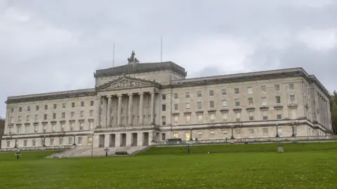 PA Media The exterior of Stormont buildings. It shows a large white building sitting on top of a hill. There are six large columns on the outside of the building and a number of windows. The sky is cloudy.