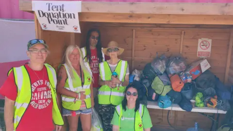 Angus Clark Two men and three women all wearing high vis jackets by a shed which is full of tents wrapped up in their bags. The men are wearing red T-shirts and one of the women is wearing a green T-shirt. There is a sign saying "Tent Donation Point" at the top of the shed.