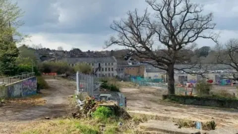 Saxonvale site in the middle of Frome looking derelict. Tree in the foreground and old empty factory buildings in the background There are bits of greenery among the paths. There are a couple of sets of railings, one of them above a wall of graffiti.