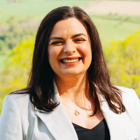 Scottish National Party A woman with shoulder-length brown hair, wearing a light grey jacket and a black top. Trees and fields can be seen in the distance.