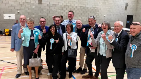 Alex Dunlop/BBC 12 men and women many wearing Reform UK turquoise rosettes with their thumbs up smile for the camera