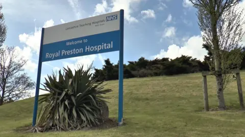 A blue and white sign reading 'Welcome to Royal Preston Hospital' in front of green bushes and and blue cloudy sky.