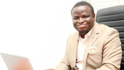 Samson Folarin A black man smiles towards the camera. He is dressed smartly in a cream suit, with pocket handkerchief and sitting in an office chair at a laptop with a telephone on the desk