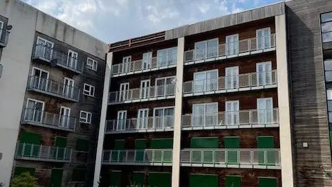 Andrew Broadhurst A block of flats. There are four levels, each with outside balconies and French doors. There is wooden cladding on some parts of the building, with other areas are covered in white stone. 