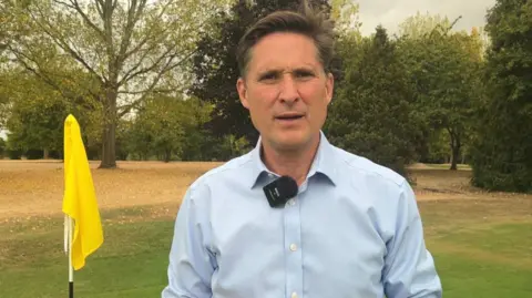 BBC/Lilah Parsons Charlie Maynard in a light blue button-down shirt stands beside a yellow golf flag on a golf green, with trees and dry fairway grass in the background. A clip-on microphone is attached to his shirt.