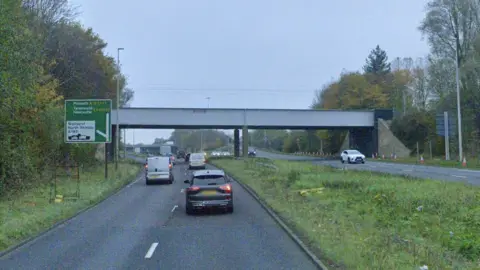 A Google Streetview screenshot of the railway bridge crossing the A19 near Howdon, North Tyneside. There are more cars on the left hand side than the right and, while there is a good amount of traffic, it is not overly busy. A green junction sign directs traffic straight on to Morpeth, Tynemouth and Newcastle, or left to Wallsend and North Shields. There is also a ferry outline on the sign, with a car and van sitting in the body of the ship to denote a vehicle ferry. The sky seems overcast and all the vehicles have their lights on.