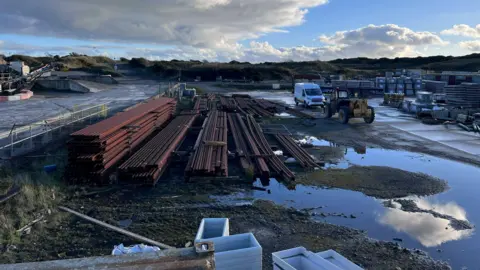 IOM GOVERNMENT Long components of a railway track piled together on the existing site, with vehicles and and other it3em stacked up on the right. There are pools of water on the ground.