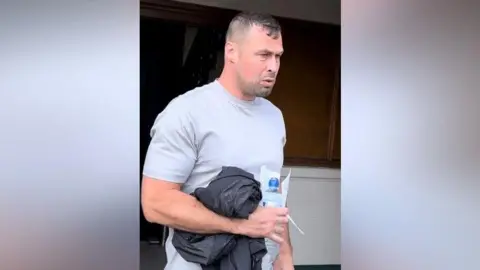 Craig McGlasson Allan Sutton, a man with short brown hair. He's wearing a grey t-shirt and holding a jacket, water bottle and sheet of paper.